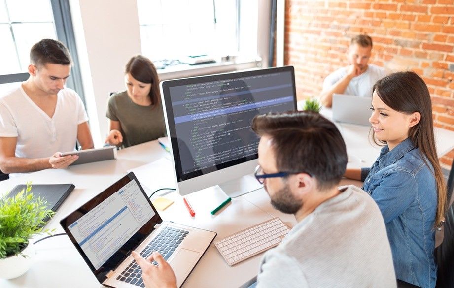 A group of five professionals collaborates around a large white desk in a bright office with an exposed brick wall. In the foreground, a man with glasses points to a laptop screen while sitting next to a smiling woman, and both look at a desktop monitor displaying code. In the background, three other colleagues work on tablets and laptops.