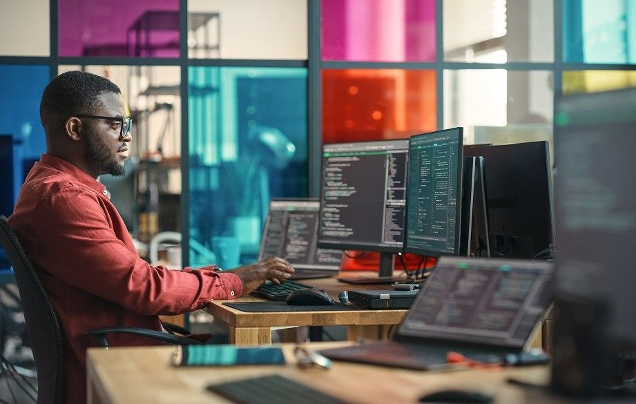 A Black man with glasses and a beard, wearing a red button-down shirt, sits at a wooden desk in a modern office, focused on writing code. He is working with two large computer monitors and a laptop, all displaying lines of programming code. The background features a contemporary glass partition with vibrant, multi-colored panels in shades of red, teal, and yellow.