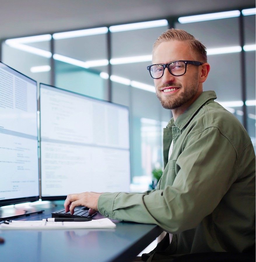 A smiling man with glasses and a short beard, wearing an olive green shirt over a white t-shirt, looks toward the camera while sitting at a sleek desk. He has a dual-monitor setup in front of him displaying technical documents or code. The office environment is bright and contemporary, with large glass partitions and soft lighting in the background.