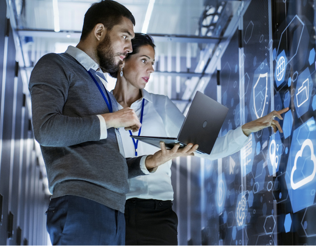 Two IT professionals stand in a server room analyzing a glowing blue digital screen with tech icons.