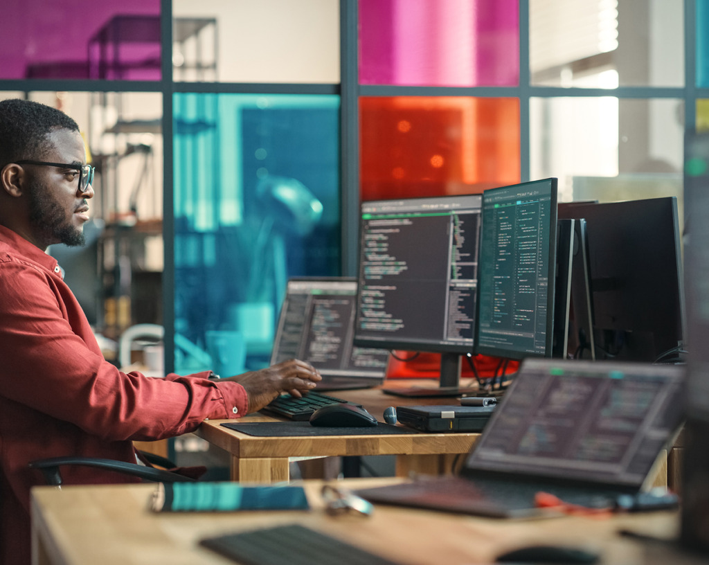 A professional software developer wearing glasses sits at a desk typing code on multiple monitors.