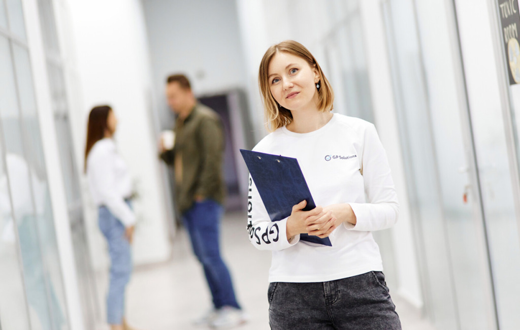 A woman wearing a white long-sleeve shirt stands in a bright office hallway holding a blue clipboard.
